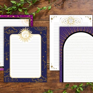 Puede incluir: Una colección de blocs de notas con temática celestial. Los blocs tienen páginas rayadas con bordes decorativos en tonos morado, azul marino y dorado. Los diseños incluyen soles, lunas, estrellas y constelaciones. La papelería está dispuesta sobre una superficie de madera.