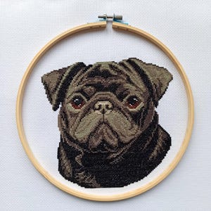 May include: Cross-stitch portrait of a pug dog with brown and black thread, framed in a wooden embroidery hoop. The pug's face is detailed with brown eyes and a wrinkled snout, set against a white fabric background.