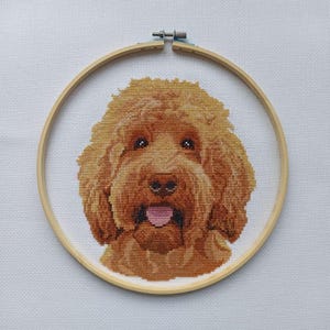 May include: A cross-stitch portrait of a golden doodle dog, framed in a wooden embroidery hoop. The dog has brown fur, dark eyes, and a pink tongue. The background is white.