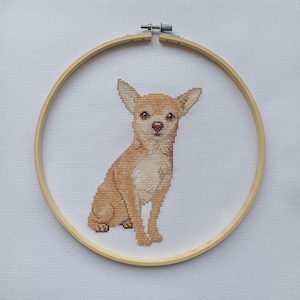Puede incluir: Bordado de punto de cruz de un perro chihuahua marrón claro sentado erguido, enmarcado en un bastidor de bordado de madera. El perro tiene orejas grandes y un pecho de color claro. El fondo es de tela blanca.