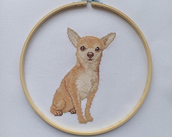 以下が含まれることがあります： 木製の刺繍枠に収められた、座っている薄茶色のチワワ犬のクロスステッチ刺繍。犬は大きな耳と明るい色の胸をしています。背景は白い布です。