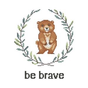 Op de afbeelding: Kruissteekontwerp met een bruine beer die zijn poten vasthoudt, omringd door een krans van groene en blauwe bladeren. De woorden "be brave" zijn in zwarte blokletters eronder gestikt.