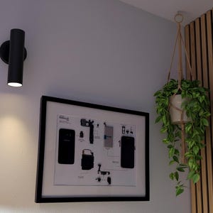 Può includere: Un'applique cilindrica nera illumina una parete bianca. Sotto, è esposto un diagramma incorniciato di un telefono Galaxy S7. Una pianta sospesa in un vaso bianco è appesa al soffitto.