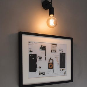 May include: A black framed print of a Google Pixel 3 phone's internal components, mounted on a gray wall. Above the print is a black, industrial-style light fixture with a glowing Edison bulb.