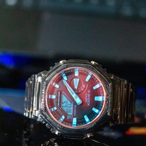 May include: A G-Shock Casio watch with a red face and a silver band. The watch displays the time 19:38. The watch has blue accents and the text "G-SHOCK CASIO" is visible.