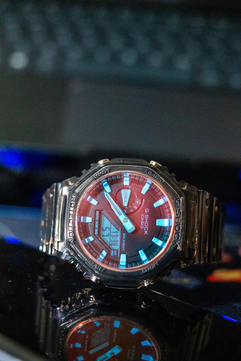May include: A G-Shock Casio watch with a red face and a silver band. The watch displays the time 19:38. The watch has blue accents and the text "G-SHOCK CASIO" is visible.