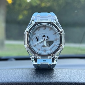 Puede incluir: Un reloj Casio G-Shock con caja y correa de resina transparente. La esfera es de color gris claro con marcadores de hora plateados, manecillas blancas y una ventana de visualización digital que muestra la fecha 1.24 y la hora 47.