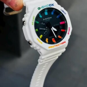 May include: White G-Shock watch with a black face and colorful accents. The watch features a white band and a multi-colored bezel with the words "G-SHOCK" and "CASIO" printed on the face. The watch has a digital display and various buttons.