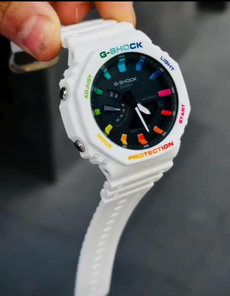 May include: White G-Shock watch with a black face and colorful accents. The watch features a white band and a multi-colored bezel with the words "G-SHOCK" and "CASIO" printed on the face. The watch has a digital display and various buttons.