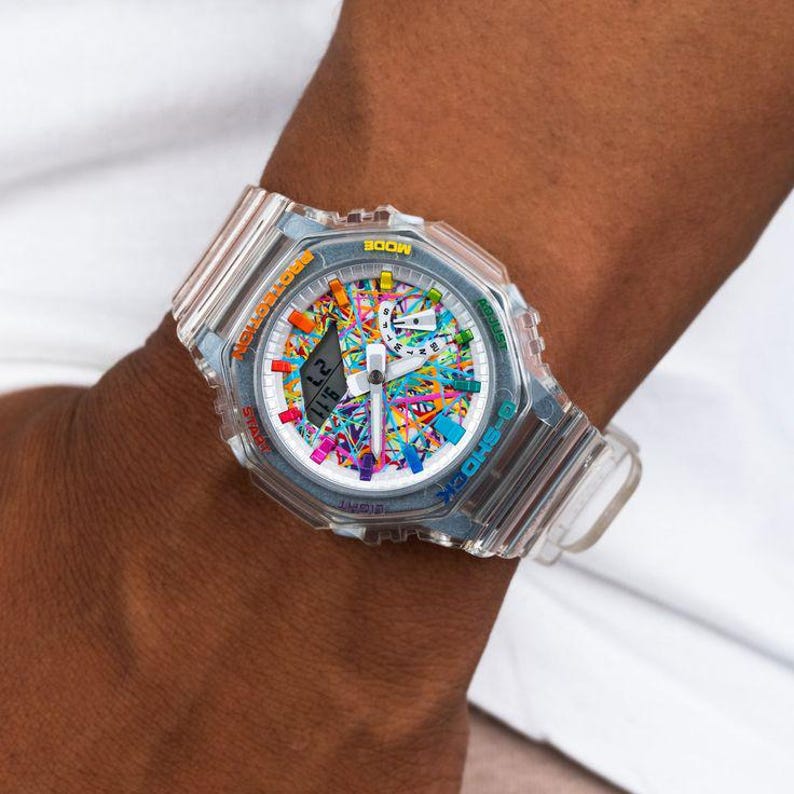 May include: A clear, transparent watch with a colorful, abstract design on the face. The watch has a digital display showing the time. The watchband is also clear. The watch is on a wrist.