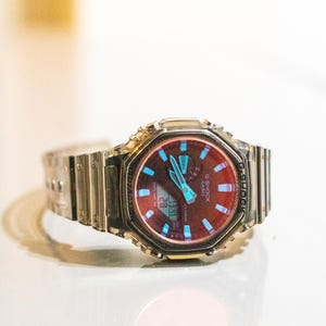 May include: A silver G-Shock watch with a red and blue face. The watch has a digital display showing the date and time. The watch band is made of silver metal.