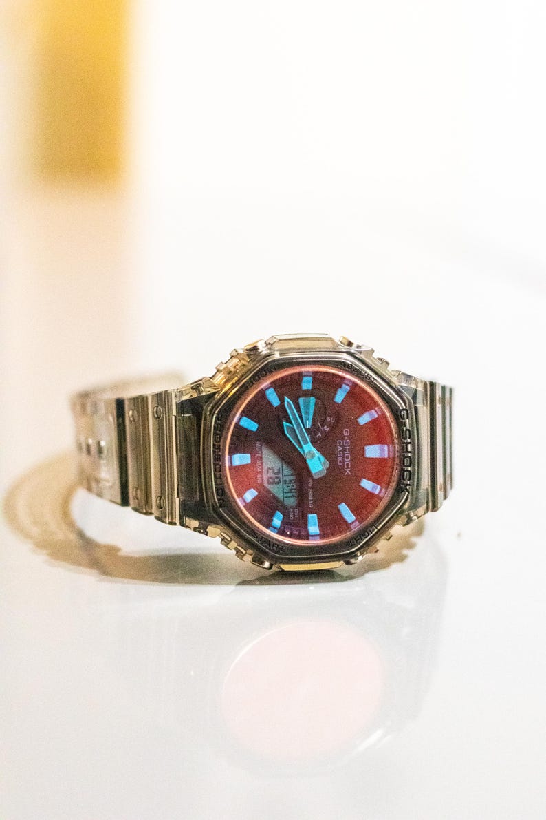 May include: A silver G-Shock watch with a red and blue face. The watch has a digital display showing the date and time. The watch band is made of silver metal.