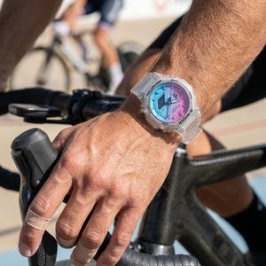 May include: A clear, transparent G-Shock watch with a gradient dial in shades of blue, pink, and purple. The watch is on a wrist, with a bicycle handlebar and a cyclist in the background. The watch has a digital display.