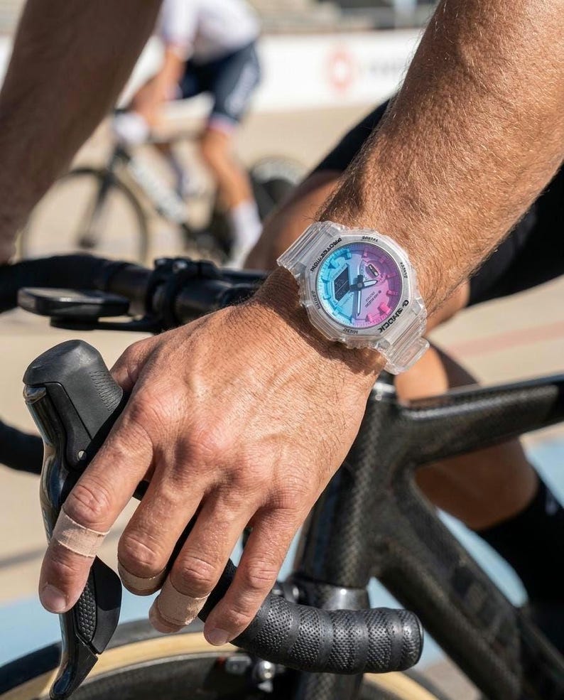 May include: A clear, transparent G-Shock watch with a gradient dial in shades of blue, pink, and purple. The watch is on a wrist, with a bicycle handlebar and a cyclist in the background. The watch has a digital display.