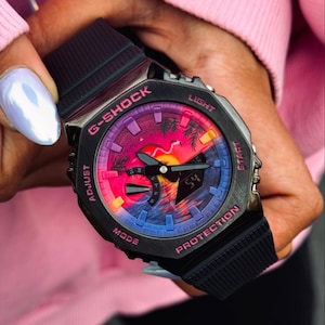 May include: Black G-Shock watch with a vibrant sunset-themed face. The watch features a black band and case with pink and purple accents. The watch face displays a colorful sunset scene with palm trees.
