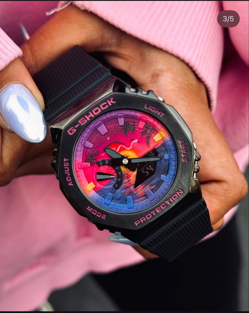 May include: Black G-Shock watch with a vibrant sunset-themed face. The watch features a black band and case with pink and purple accents. The watch face displays a colorful sunset scene with palm trees.