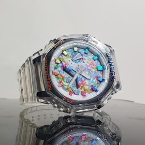 May include: A clear G-Shock watch with a colorful face featuring a mix of colors. The watch has a clear band and a digital display. The watch has the words "Adjust", "Protection", "Start", and "Mode" on the bezel.