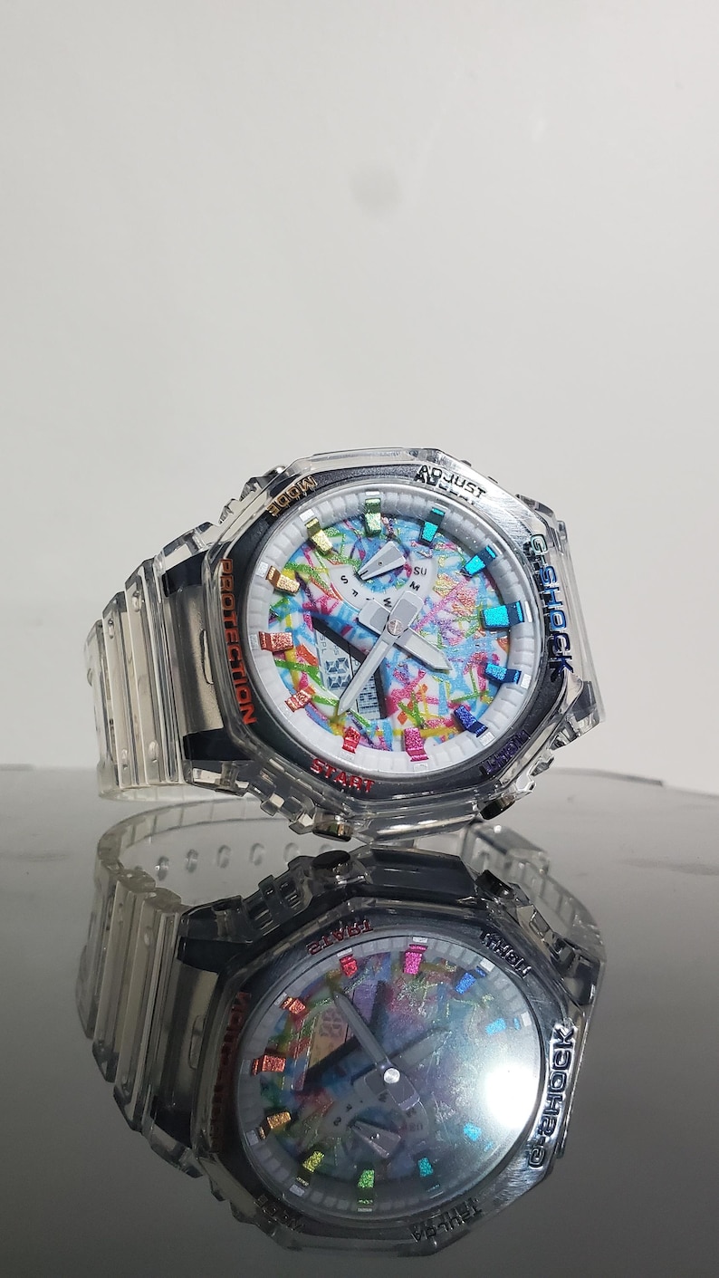 May include: A clear G-Shock watch with a colorful face featuring a mix of colors. The watch has a clear band and a digital display. The watch has the words "Adjust", "Protection", "Start", and "Mode" on the bezel.