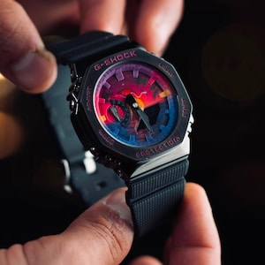 May include: Black G-Shock watch with a colorful sunset design on the face. The watch has a black band and a silver bezel. The watch face features the words "G-SHOCK" and "PROTECTION".