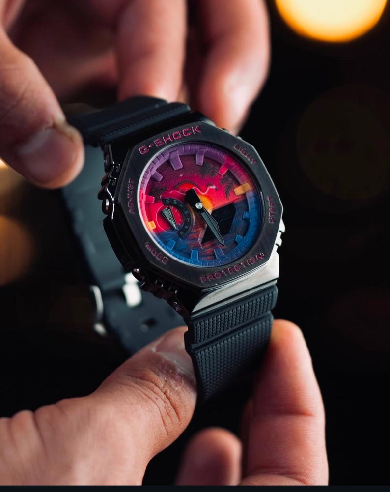 May include: Black G-Shock watch with a colorful sunset design on the face. The watch has a black band and a silver bezel. The watch face features the words "G-SHOCK" and "PROTECTION".