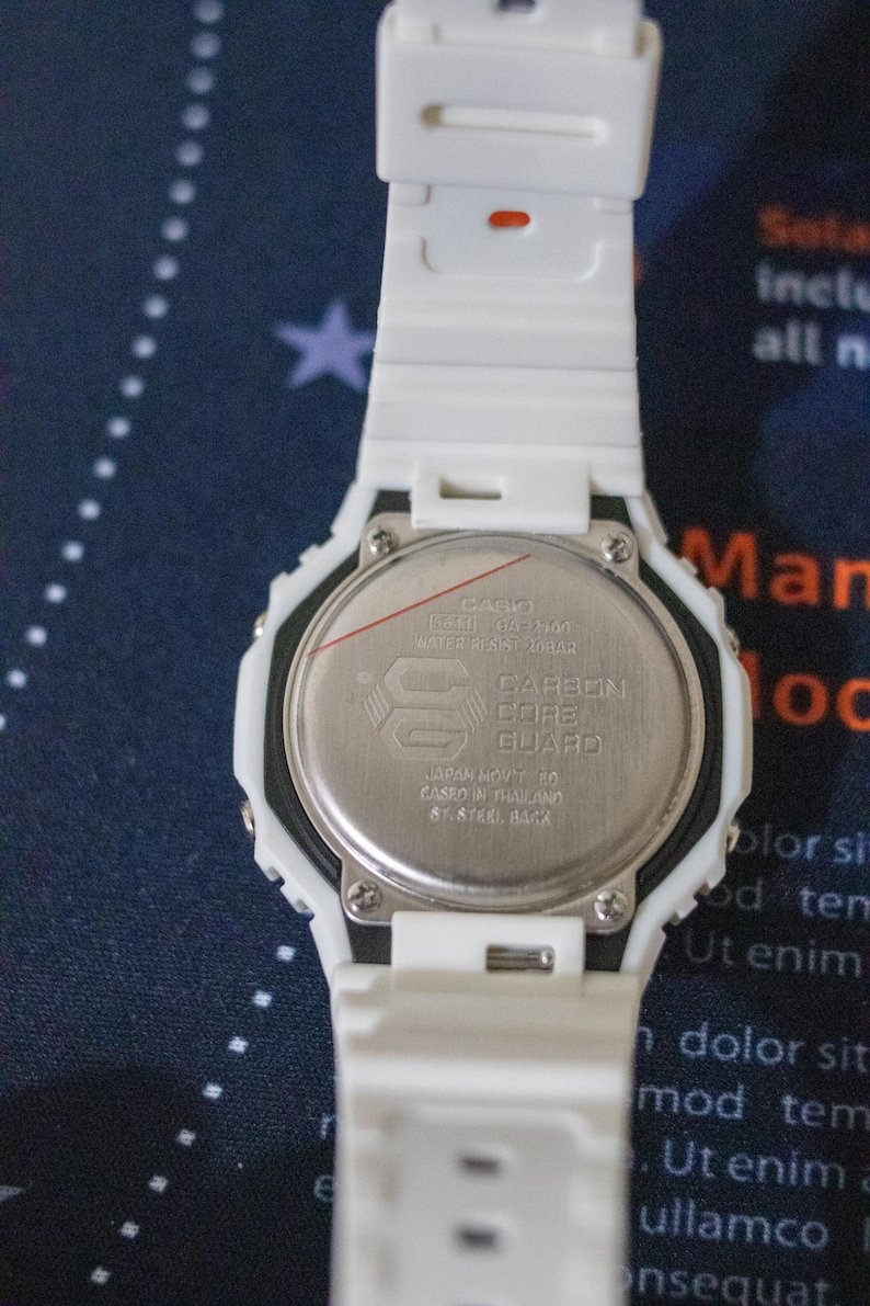 May include: A white digital watch with a white band. The watch face is silver with black text and a logo. The back of the watch has text that reads "CARBON CORE GUARD".