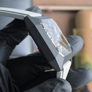 May include: A close-up of a contemporary wristwatch featuring a geometric black case and a transparent face. The watch has a white strap and is held by a person wearing black gloves. The watch face reveals intricate internal mechanisms.