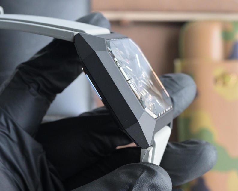 May include: A close-up of a contemporary wristwatch featuring a geometric black case and a transparent face. The watch has a white strap and is held by a person wearing black gloves. The watch face reveals intricate internal mechanisms.