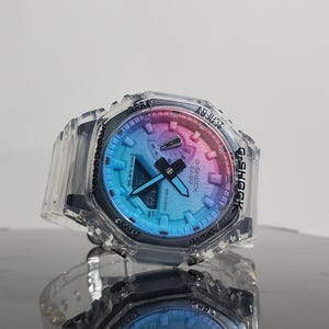 Puede incluir: Un reloj G-Shock transparente con una esfera multicolor, que muestra la hora 1:23. El reloj tiene una caja protectora transparente y una correa negra. El reloj se refleja en la superficie de abajo.