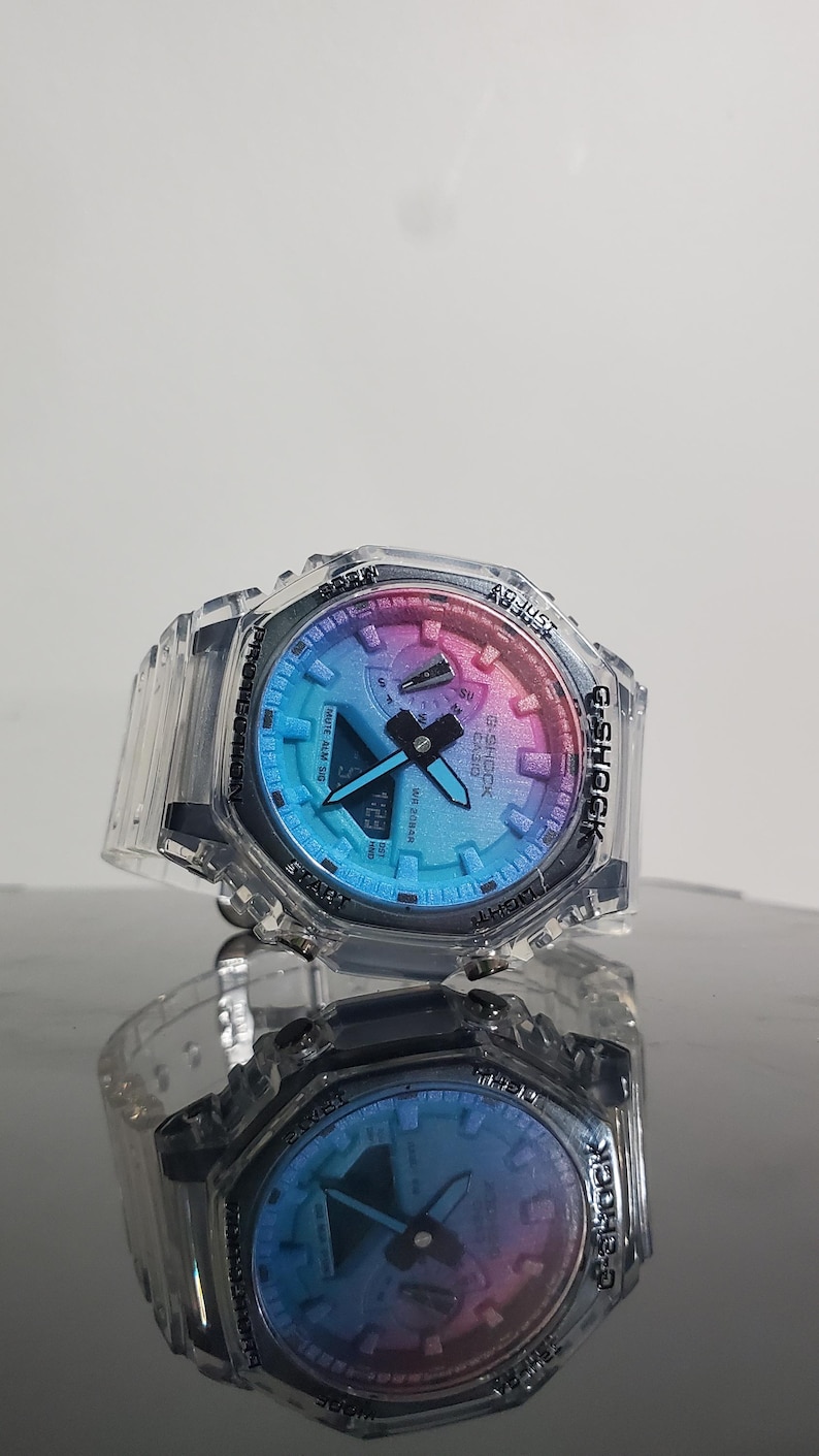 May include: A clear G-Shock watch with a multi-colored face, displaying the time as 1:23. The watch has a clear protective case and a black band. The watch is reflected in the surface below.