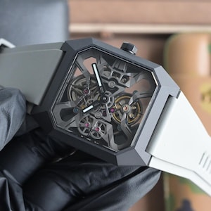 May include: A contemporary wristwatch featuring a geometric black case and a transparent face showcasing the complex internal mechanisms. The watch has a grey strap and a black crown. The watch face displays silver and gold gears and ruby-coloured jewels.