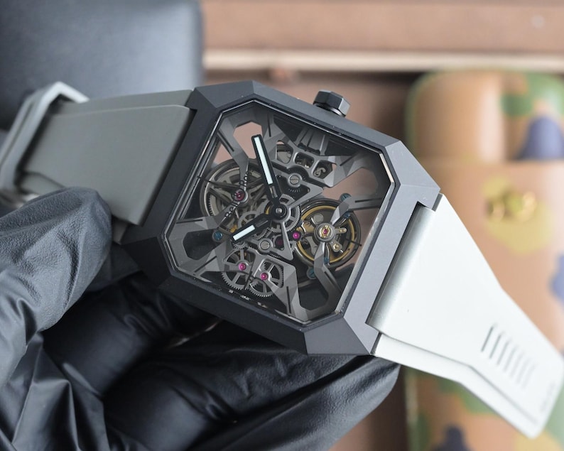 May include: A contemporary wristwatch featuring a geometric black case and a transparent face showcasing the complex internal mechanisms. The watch has a grey strap and a black crown. The watch face displays silver and gold gears and ruby-coloured jewels.