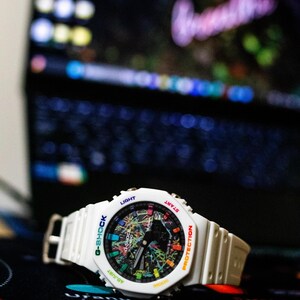 May include: White G-Shock watch with a colorful, abstract design on the face. The watch has a white band and a digital display. The watch face includes the words "G-SHOCK", "LIGHT", "START", "PROTECTION", and "ADJUST".