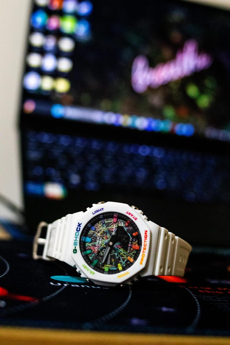 May include: White G-Shock watch with a colorful, abstract design on the face. The watch has a white band and a digital display. The watch face includes the words "G-SHOCK", "LIGHT", "START", "PROTECTION", and "ADJUST".
