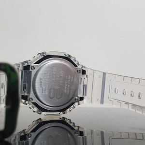 May include: A clear, transparent Casio G-Shock watch with a silver-tone face and band. The watch features a digital display and a carbon core guard structure. The watch is water resistant.