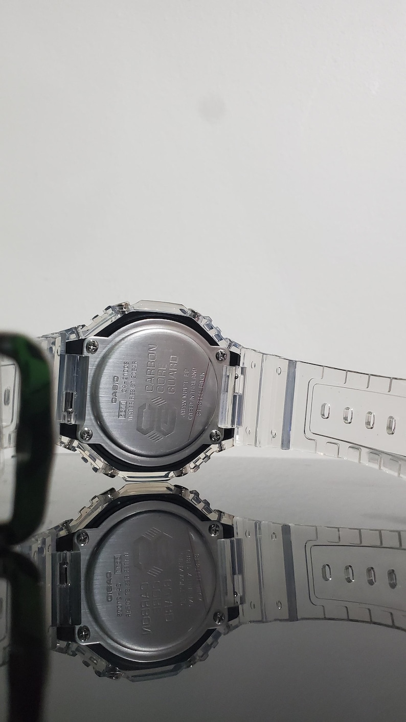 May include: A clear, transparent Casio G-Shock watch with a silver-tone face and band. The watch features a digital display and a carbon core guard structure. The watch is water resistant.