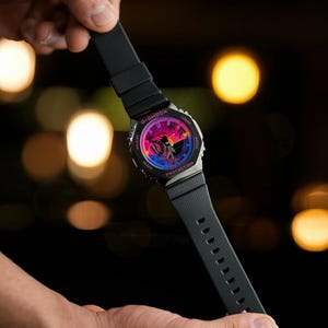 May include: Black G-Shock watch with a colorful dial featuring pink, purple, and blue hues. The watch has a black strap and a silver-tone case. The watch is held against a blurred background with bokeh lights.