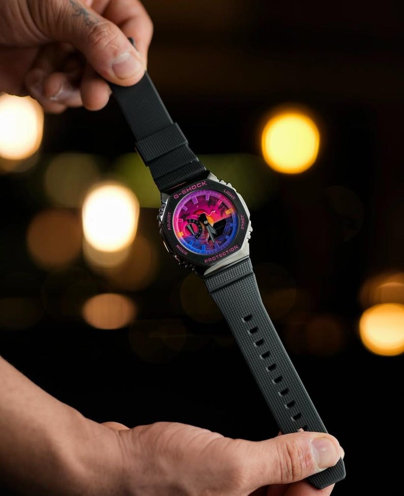 May include: Black G-Shock watch with a colorful dial featuring pink, purple, and blue hues. The watch has a black strap and a silver-tone case. The watch is held against a blurred background with bokeh lights.