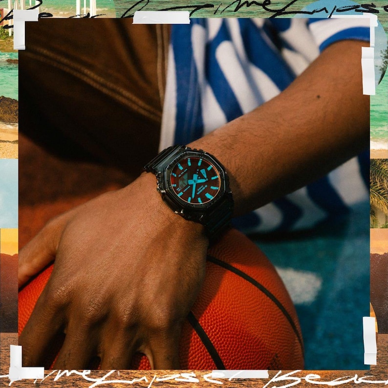 May include: Black wristwatch with a square face and a black band. The watch face has blue and red accents. The watch is on a person's wrist, with a basketball in the background.