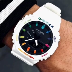 May include: White G-Shock watch with a black face and colorful accents. The watch features the text "G-SHOCK" in green and blue, and "CASIO" in white. The watch has a digital display showing the time. The watch is on a wrist.