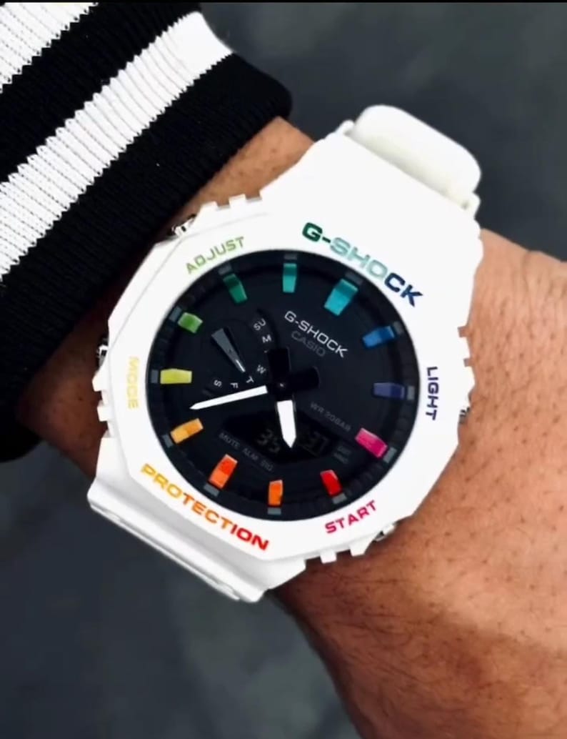 May include: White G-Shock watch with a black face and colorful accents. The watch features the text "G-SHOCK" in green and blue, and "CASIO" in white. The watch has a digital display showing the time. The watch is on a wrist.