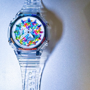 May include: Clear G-Shock watch with a colorful abstract design on the face. The watch has a clear band and case, with the words "PROTECTION" and "START" visible. The watch face features a mix of colors.