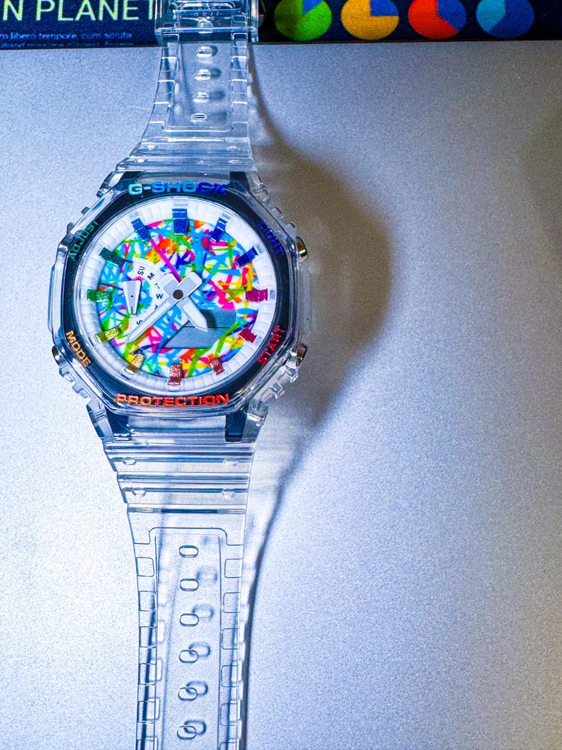 May include: Clear G-Shock watch with a colorful abstract design on the face. The watch has a clear band and case, with the words "PROTECTION" and "START" visible. The watch face features a mix of colors.