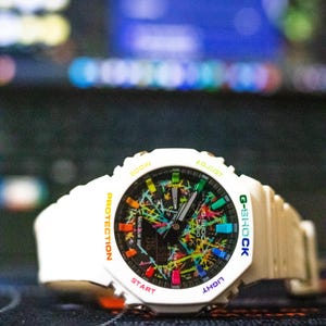 May include: White G-Shock digital watch with a colorful splatter design on the face. The watch has a white band and a black digital display. The watch face includes the text "G-SHOCK", "PROTECTION", "LIGHT", "START", and "ADJUST".