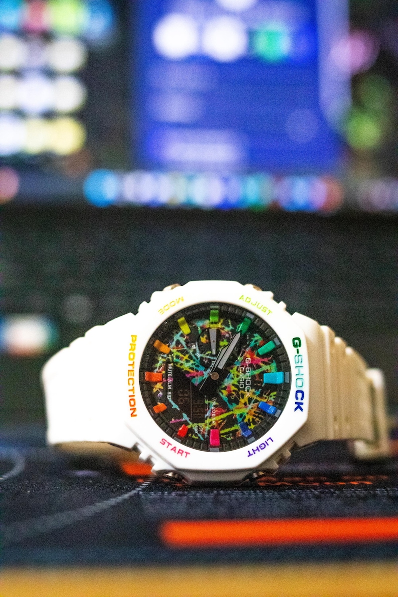 May include: White G-Shock digital watch with a colorful splatter design on the face. The watch has a white band and a black digital display. The watch face includes the text "G-SHOCK", "PROTECTION", "LIGHT", "START", and "ADJUST".