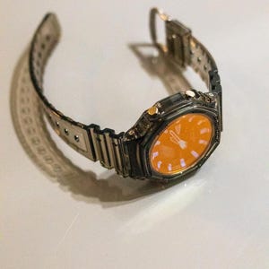 May include: A silver-toned wristwatch with an orange face. The watch has a rectangular case and a transparent band. The watch is displayed on a white surface. The watch band is approximately 9 inches long.