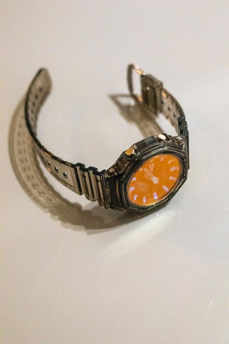 May include: A silver-toned wristwatch with an orange face. The watch has a rectangular case and a transparent band. The watch is displayed on a white surface. The watch band is approximately 9 inches long.