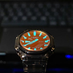 May include: A close-up of a G-Shock watch with a brown face and blue accents. The watch has a stainless steel band and a black bezel. The watch face displays the G-Shock and Casio logos.