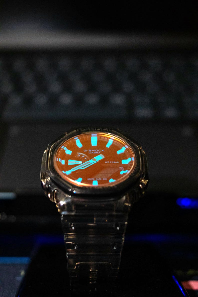 May include: A close-up of a G-Shock watch with a brown face and blue accents. The watch has a stainless steel band and a black bezel. The watch face displays the G-Shock and Casio logos.
