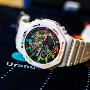 May include: White G-Shock watch with a colorful, abstract design on the face. The watch features a white band and a black dial with multi-colored accents. The watch has the words "G-SHOCK", "LIGHT", "START", "PROTECTION", "ADJUST", and "MODE" on the watch face.