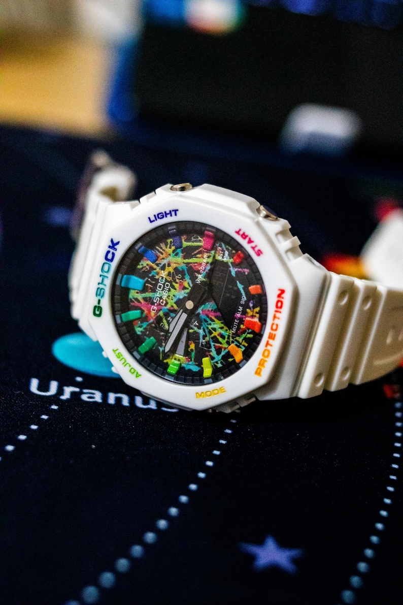 May include: White G-Shock watch with a colorful, abstract design on the face. The watch features a white band and a black dial with multi-colored accents. The watch has the words "G-SHOCK", "LIGHT", "START", "PROTECTION", "ADJUST", and "MODE" on the watch face.